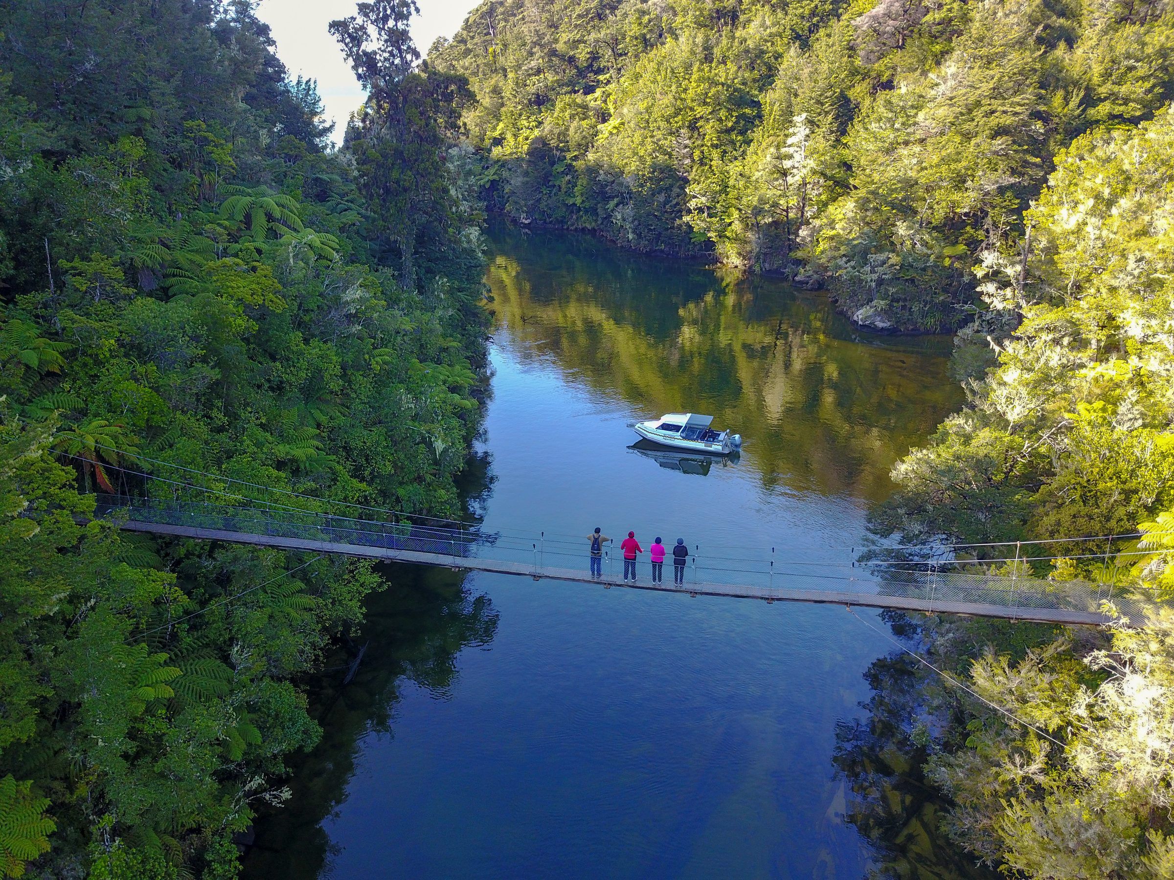 Walkers on a swing bridge above the Falls River, a boat moored in the green water below