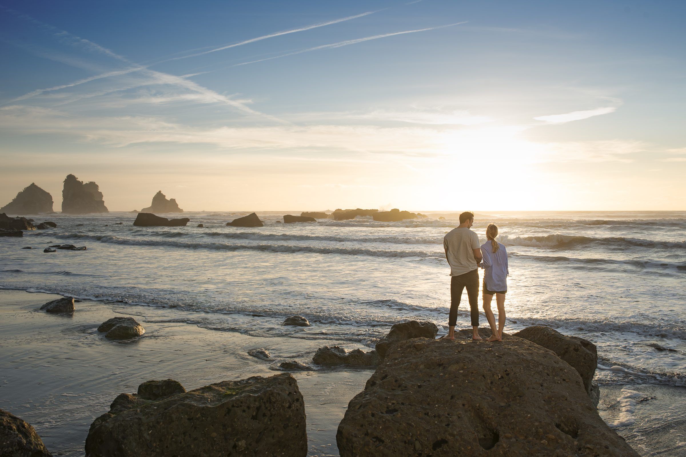 Two people stand on rocks at Rapahoe as the sun sets over the sea
