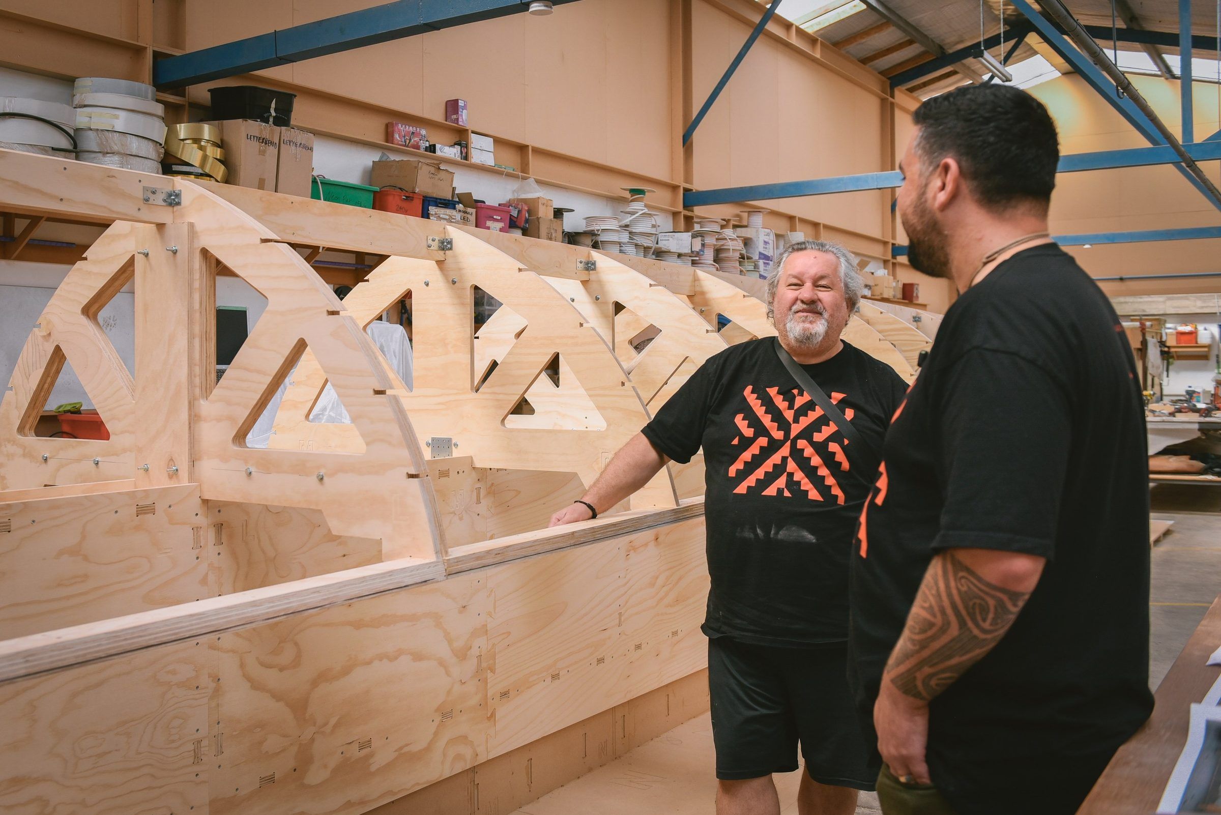 Two makers talk beside the timber frame of the waka