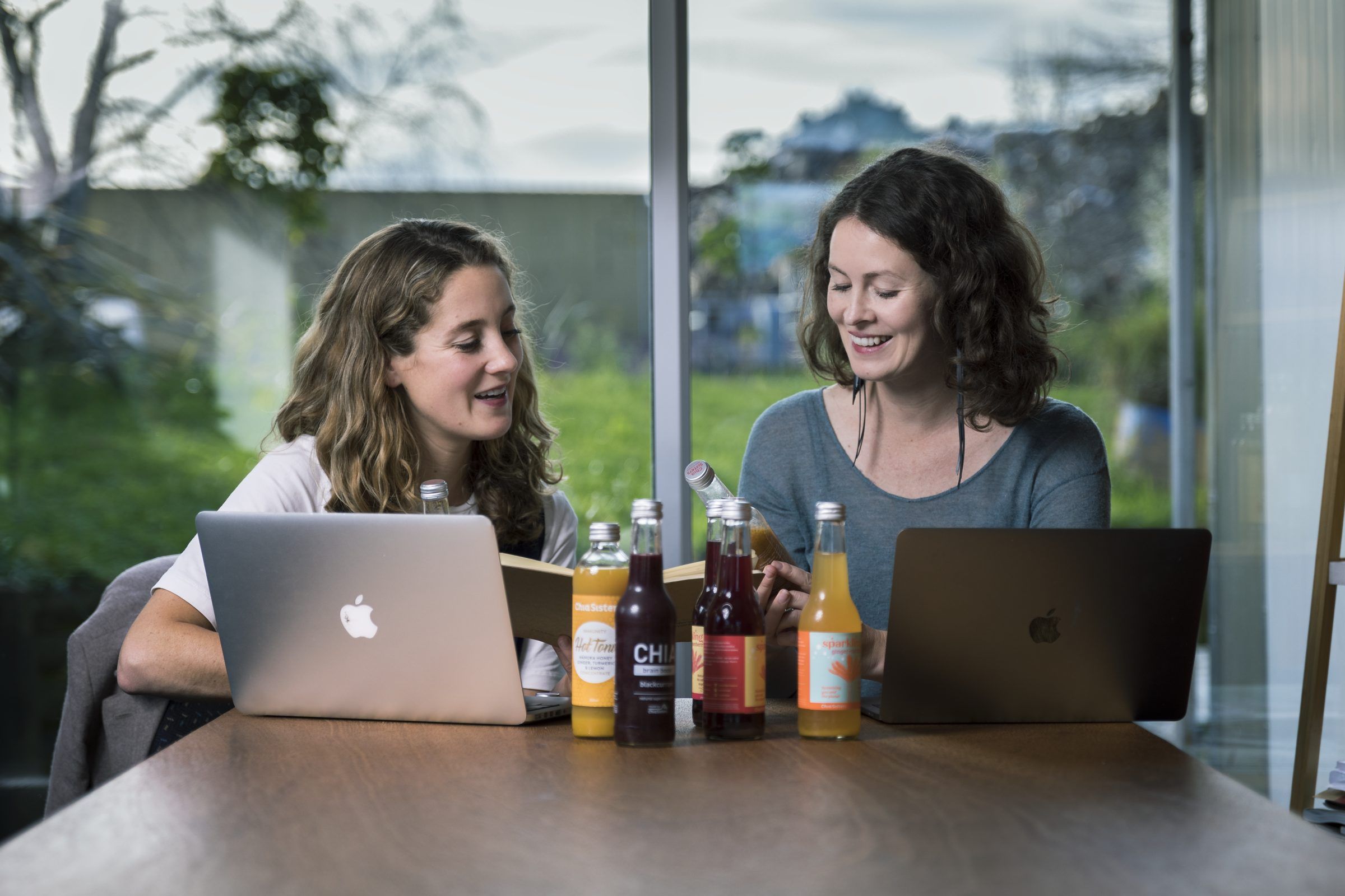 Two women working at laptops at a table with bottles of juice