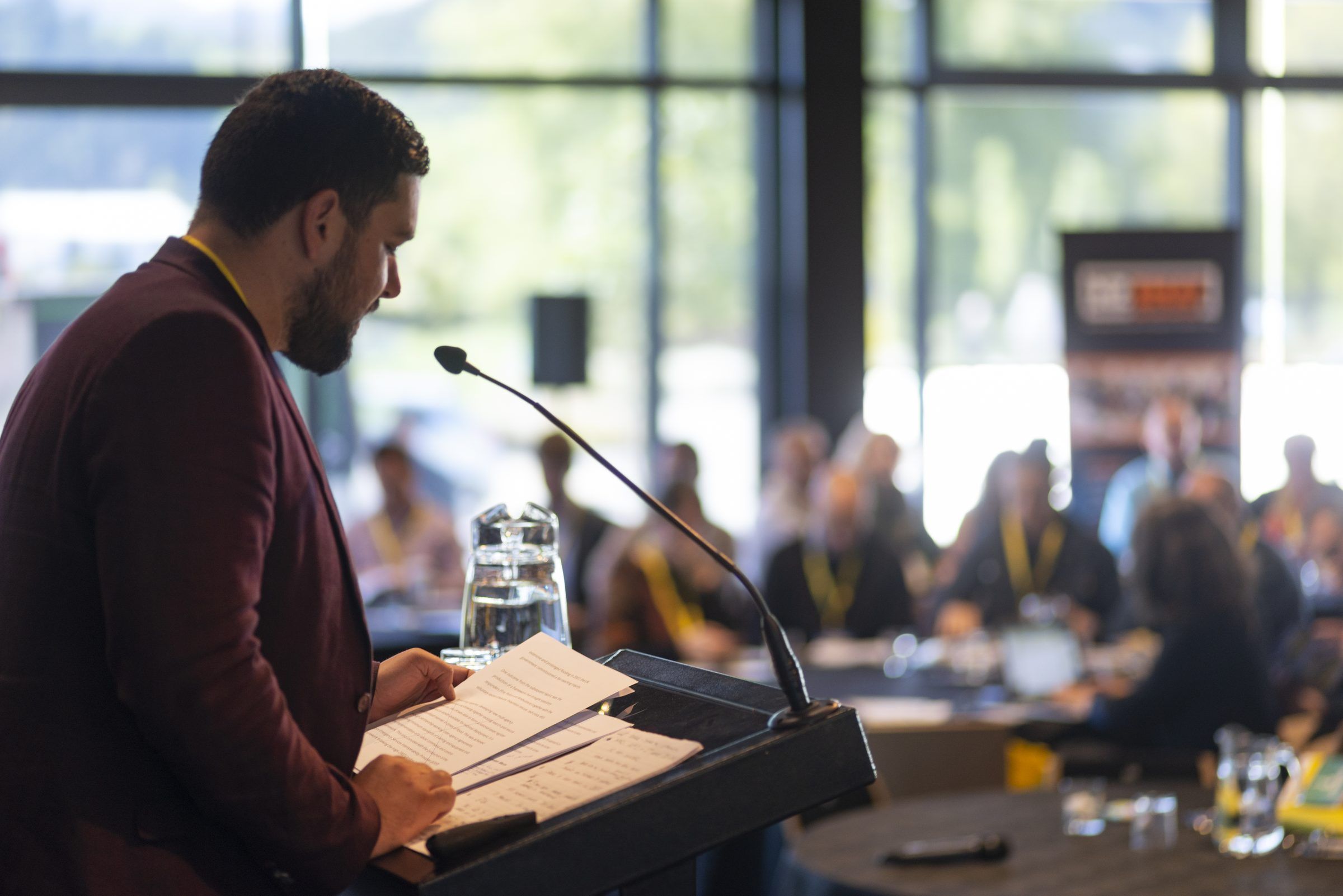 A presenter reads from a lectern to a room of people at tables