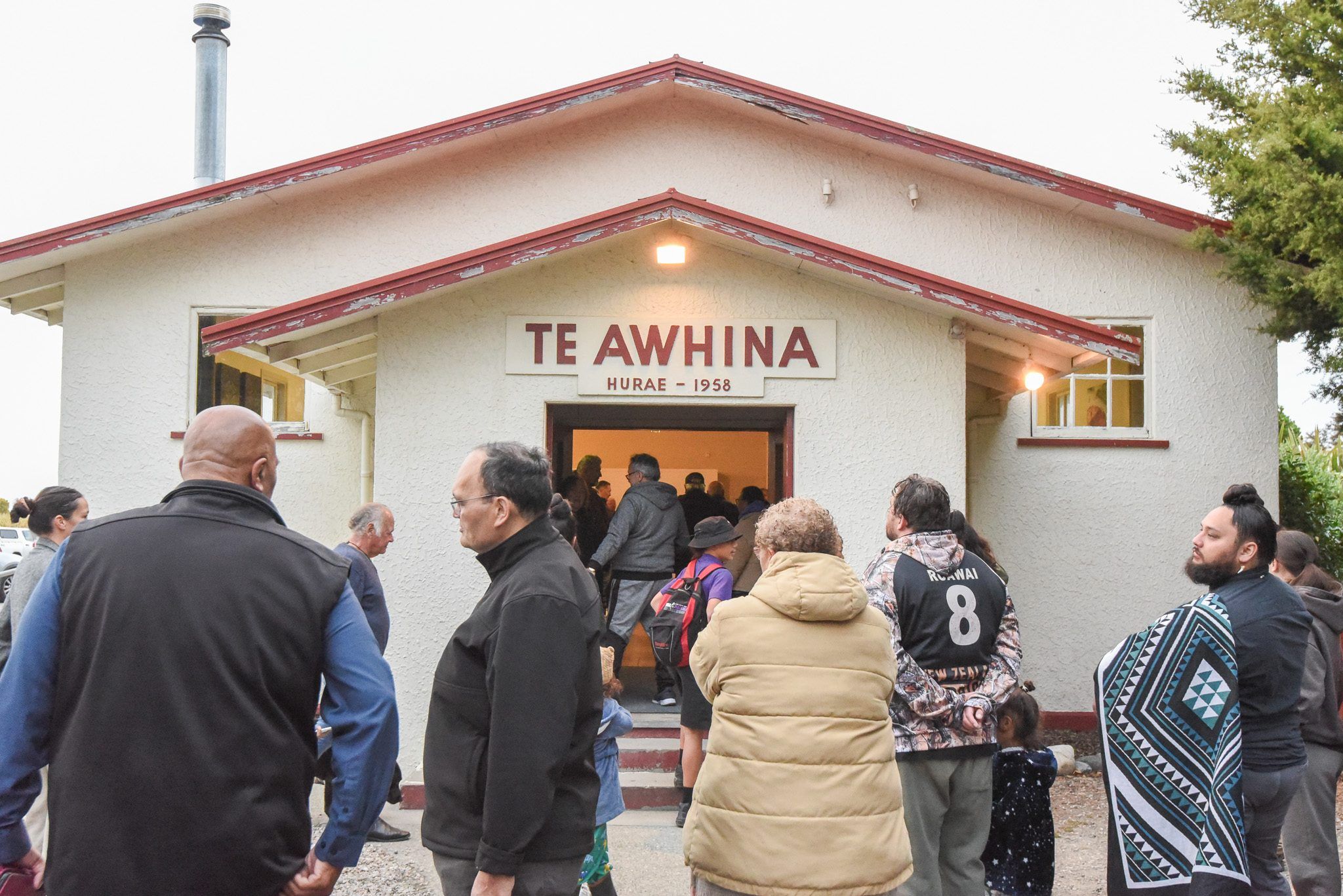 People gather at the lit entrance of the Te Awhina hall