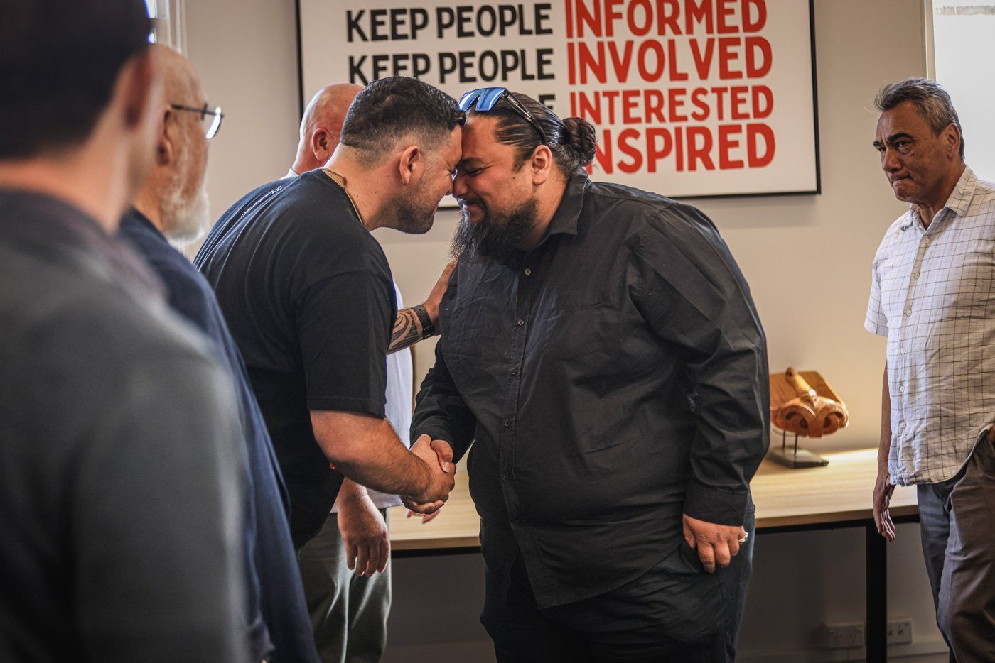 Two men share a hongi beneath a poster reading keep people informed, involved, interested, inspired